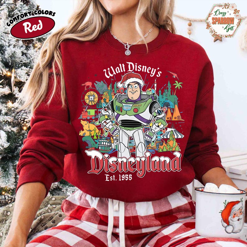 K&ouml;nnte beinhalten: Ein rotes Comfort Colors Sweatshirt mit Buzz Lightyear in einem Weihnachtsmannhut und der Aufschrift "Walt Disney's Disneyland Est. 1955". Die Grafik enth&auml;lt Disneyland-Symbole. Das Sweatshirt wird mit einer rot-wei&szlig; karierten Pyjamahose getragen.