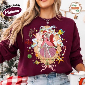 May include: Maroon sweatshirt featuring a festive illustration of a princess in a pink dress, surrounded by holiday decorations. The design includes a gold frame, lights, and sea creatures. The text "Gildan Colors Maroon" is in the upper left corner.