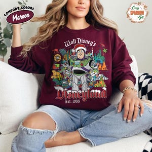K&ouml;nnte beinhalten: Burgunderfarbenes Sweatshirt mit einem Aufdruck von Buzz Lightyear mit Weihnachtsm&uuml;tze und der Aufschrift "Walt Disney's Disneyland Est. 1955". Das Design umfasst verschiedene Disneyland-Parkelemente. Das Sweatshirt ist von der Marke Comfort Colors.