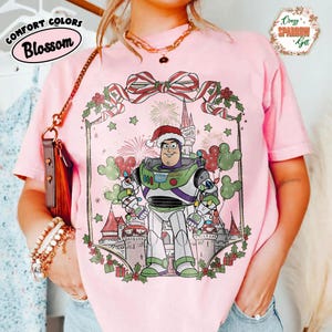 May include: Pink t-shirt featuring a graphic of Buzz Lightyear in a Santa hat, surrounded by Christmas decorations and a castle. The shirt has the words "Comfort Colors Blossom" and "Crazy Sparrow Gift".
