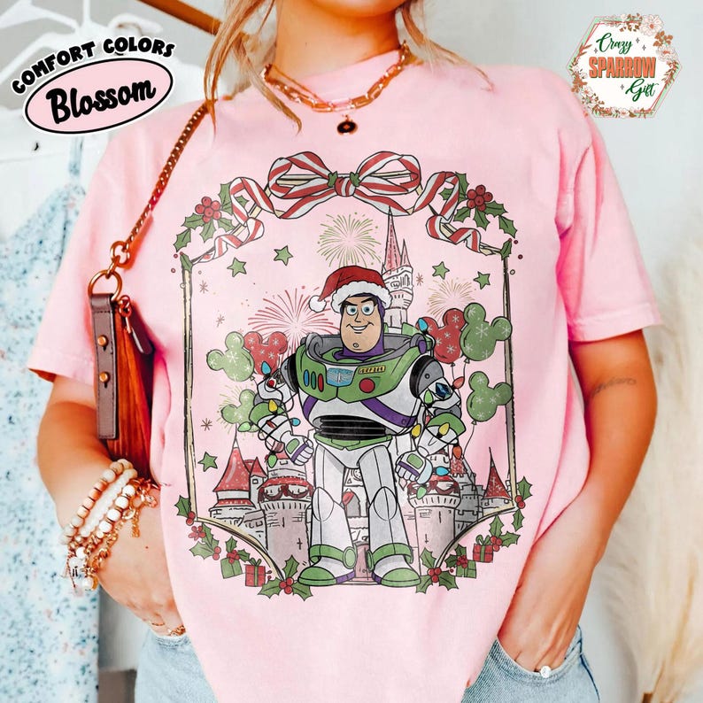 May include: Pink t-shirt featuring a graphic of Buzz Lightyear in a Santa hat, surrounded by Christmas decorations and a castle. The shirt has the words "Comfort Colors Blossom" and "Crazy Sparrow Gift".