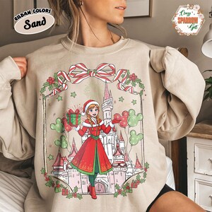 Może przedstawiać: Beżowa bluza z uroczystą ilustracją. Projekt zawiera postać w czerwono-zielonym stroju, trzymającą prezent i balony, z zamkiem w tle. Obraz jest obramowany kokardą z cukrowej laski i ostrokrzewem. Widoczny jest tekst "GILDAN COLORS Sand".