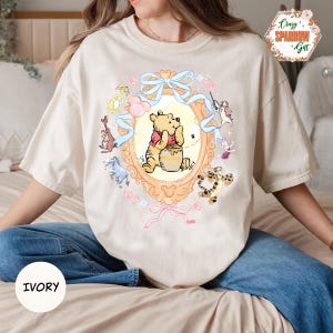 Puede incluir: Camiseta de color marfil con una ilustración enmarcada de Winnie the Pooh y sus amigos. El diseño incluye un marco dorado con lazos azules y detalles en rosa. El texto "IVORY" está en la parte inferior de la camiseta.