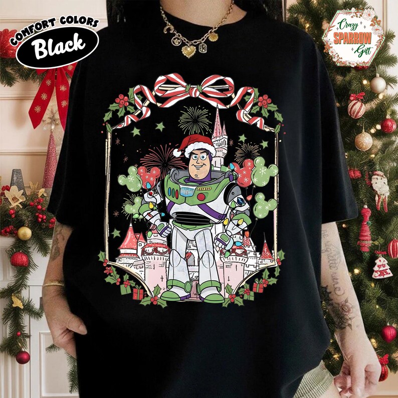 May include: Black t-shirt featuring a festive Buzz Lightyear design wearing a Santa hat, standing in front of a castle with fireworks and Mickey Mouse-shaped ornaments. The shirt has the words "Comfort Colors Black" in a white oval.