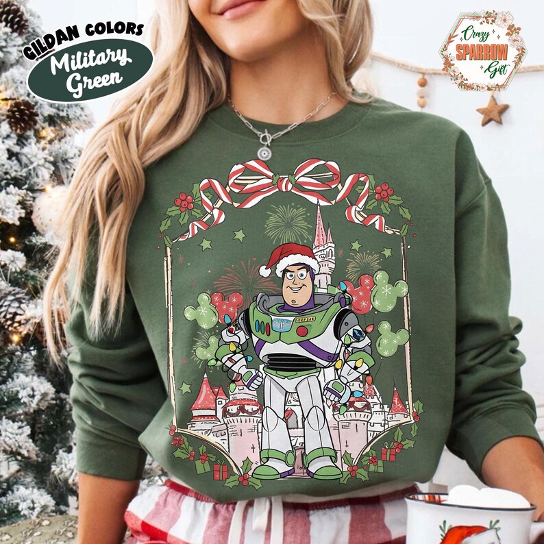 May include: Military green sweatshirt featuring a graphic of Buzz Lightyear in a Santa hat, surrounded by a Christmas-themed design with a castle and fireworks. The sweatshirt has the text "GILDAN COLORS Military Green".