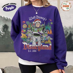 K&ouml;nnte beinhalten: Lila Comfort Colors Sweatshirt mit einer Grafik von Buzz Lightyear mit Weihnachtsm&uuml;tze, umgeben von Disneyland-Bildern. Der Text "Walt Disney's Disneyland Est. 1955" ist in Rot und Wei&szlig; dargestellt. Ein rundes Comfort Colors-Logo befindet sich oben links.