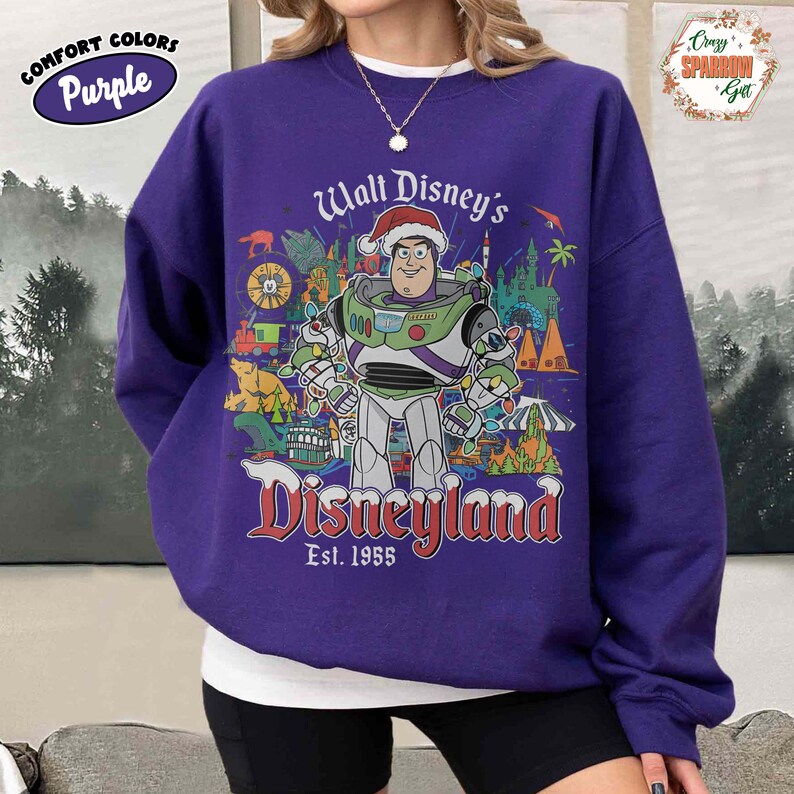 K&ouml;nnte beinhalten: Lila Comfort Colors Sweatshirt mit einer Grafik von Buzz Lightyear mit Weihnachtsm&uuml;tze, umgeben von Disneyland-Bildern. Der Text "Walt Disney's Disneyland Est. 1955" ist in Rot und Wei&szlig; dargestellt. Ein rundes Comfort Colors-Logo befindet sich oben links.
