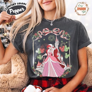 May include: Dark grey Comfort Colors t-shirt with a Christmas graphic. The design features a princess in a pink dress, a castle, fireworks, and Mickey Mouse silhouettes. The shirt has the text "Pepper" and "Crazy Sparrow Get".