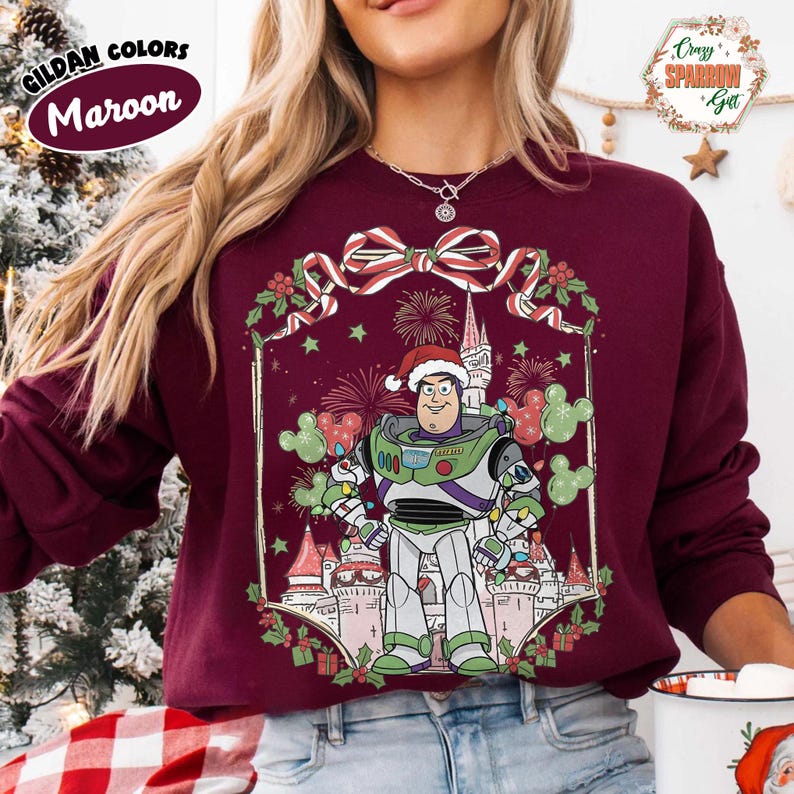 May include: Maroon sweatshirt featuring a festive Buzz Lightyear design. The graphic includes a castle, fireworks, and Mickey Mouse-shaped balloons. The text "Cozy Sparrow Get" is visible. The sweatshirt is a Gildan color.