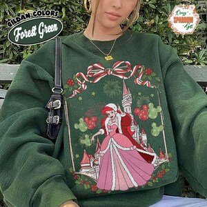 May include: Forest green sweatshirt with a Christmas design, including a princess, castle, and Mickey Mouse shapes. The sweatshirt has a crew neck and long sleeves. The text "GILDAN COLORS Forest Green" is visible on the top left.