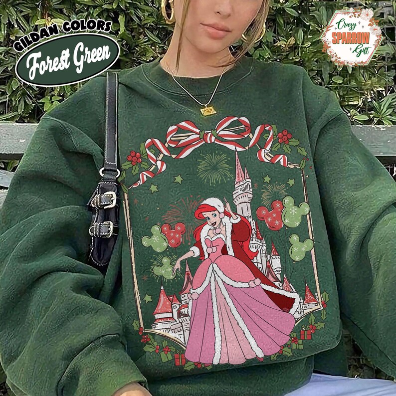 May include: Forest green sweatshirt with a Christmas design, including a princess, castle, and Mickey Mouse shapes. The sweatshirt has a crew neck and long sleeves. The text "GILDAN COLORS Forest Green" is visible on the top left.
