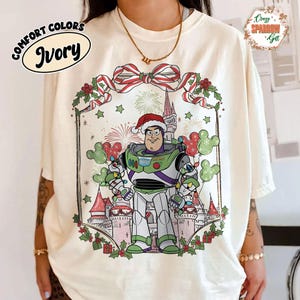 May include: Ivory Comfort Colors t-shirt featuring a festive Buzz Lightyear graphic wearing a Santa hat, surrounded by Christmas lights and a castle scene. The shirt includes the text "Comfort Colors Ivory" in a circular design.