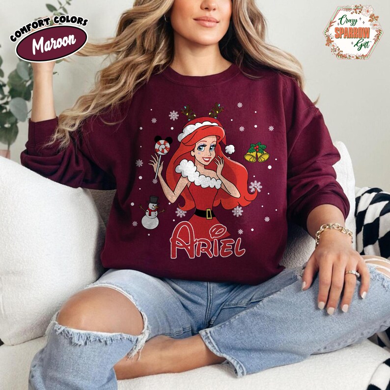 May include: Maroon sweatshirt featuring a festive Ariel graphic. The design includes Ariel in a Santa hat, holding a candy cane, with snowflakes and a snowman. The word "Ariel" is written in a script font below the graphic.
