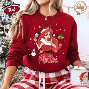 May include: A red sweatshirt featuring a Christmas-themed Ariel design. The graphic includes Ariel in a red dress, snowflakes, a snowman, and the name "Ariel". The sweatshirt has long sleeves and a crew neck, perfect for the holiday season.
