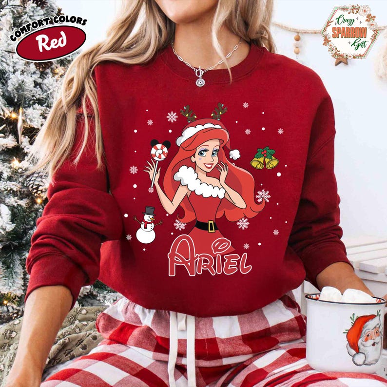 May include: A red sweatshirt featuring a Christmas-themed Ariel design. The graphic includes Ariel in a red dress, snowflakes, a snowman, and the name "Ariel". The sweatshirt has long sleeves and a crew neck, perfect for the holiday season.