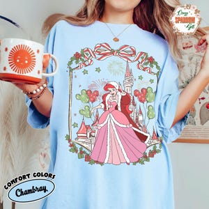 May include: Pale blue Comfort Colors Chambray t-shirt with a festive illustration. The design features a princess in a pink dress, a castle, Mickey Mouse balloons, and a Christmas bow. The shirt also has the text "Crazy Sparrow Gift".
