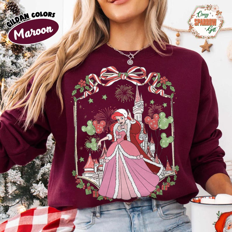 May include: Maroon sweatshirt with a princess illustration in a pink gown and a castle, Mickey Mouse balloons and fireworks. The sweatshirt has the text "Gildan Colors Maroon" on the upper left.