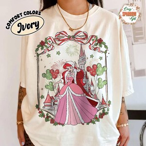May include: Ivory Comfort Colors t-shirt with a festive illustration of a princess in a pink gown and red cape, surrounded by a castle, Mickey Mouse balloons, and holiday decorations. The shirt has the text "Comfort Colors Ivory" and "Crazy Sparrow Gift".