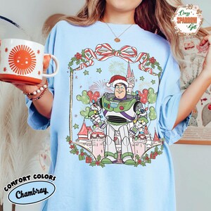 May include: Light blue Comfort Colors Chambray t-shirt featuring a festive Buzz Lightyear graphic wearing a Santa hat. The design includes a castle, fireworks, and Mickey Mouse-shaped balloons, with the text "Comfort Colors Chambray".