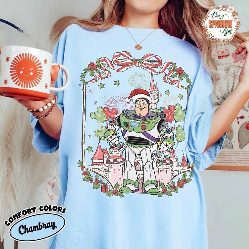 May include: Light blue Comfort Colors Chambray t-shirt featuring a festive Buzz Lightyear graphic wearing a Santa hat. The design includes a castle, fireworks, and Mickey Mouse-shaped balloons, with the text "Comfort Colors Chambray".