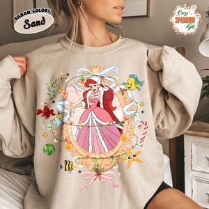 May include: Sand-colored sweatshirt featuring a festive illustration of a princess in a pink dress, surrounded by Christmas lights and sea creatures. The sweatshirt has the text "GILDAN COLORS Sand" in the upper left corner.