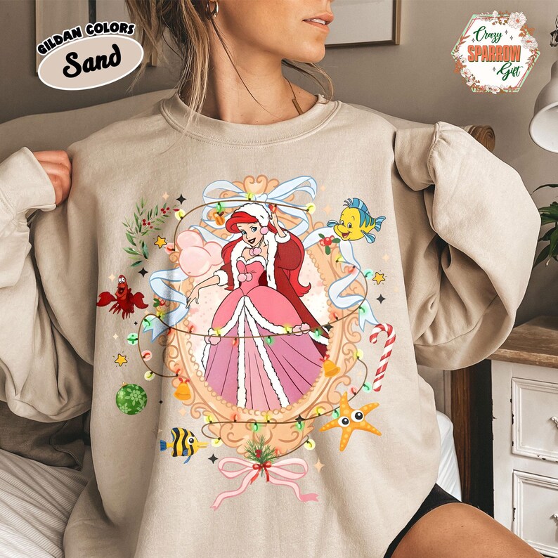 May include: Sand-colored sweatshirt featuring a festive illustration of a princess in a pink dress, surrounded by Christmas lights and sea creatures. The sweatshirt has the text "GILDAN COLORS Sand" in the upper left corner.