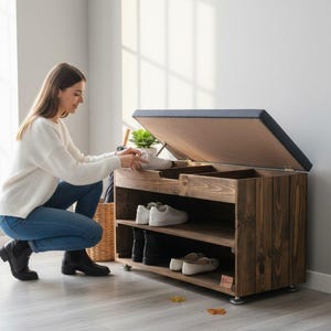 Könnte beinhalten: Eine dunkelbraune Schuhaufbewahrungsbank aus Holz mit zwei offenen Regalen und einem aufklappbaren Deckel mit dunkelgrauem Polstersitz. Das Möbelstück hat kleine Metallfüße und steht auf einem hellen Boden in der Nähe eines Fensters.
