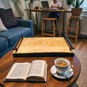 May include: A wooden serving tray with black handles sits on a round, dark wood coffee table. An open book and a cup of coffee on a saucer are also on the table. A person is working on a laptop at a desk in the background.
