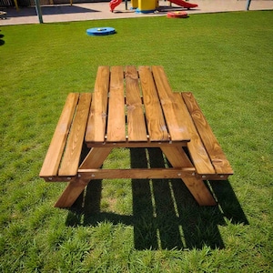 以下が含まれることがあります： 木製のピクニックテーブルとベンチ。テーブルは明るい茶色の木材でできており、クラシックなデザインです。テーブルは緑の芝生の上に置かれ、背景には遊び場が見えます。