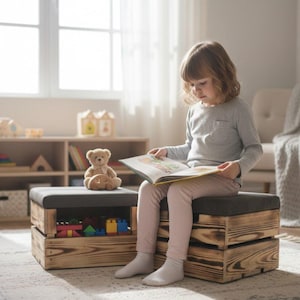 May include: A child sits on a wooden storage ottoman, reading a book. The ottoman has a gray cushioned top and is made of rustic, stained wood. A teddy bear sits on top of the ottoman. Toys are visible inside the storage space.