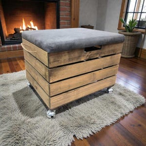 Peut inclure: Un pouf de rangement en bois avec un dessus rembourré gris. La base en forme de caisse en bois est en bois teinté et possède une poignée sur le côté. Le pouf repose sur quatre roulettes argentées et est placé sur un tapis gris duveteux.