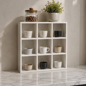 May include: A white wooden square shelving unit with nine individual compartments. Inside the cubbies are various small ceramic cups in white, grey, black, and beige tones. A glass jar with a wooden lid and a small green plant sit on top.
