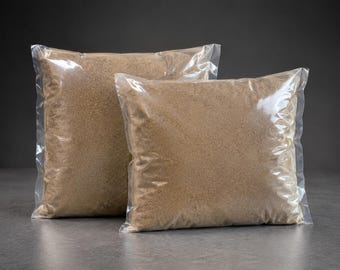 May include: Two square, clear plastic bags filled with a fine, light brown granular substance. The bags are sealed and positioned one slightly behind the other, showcasing the uniform texture and sandy color of the contents inside each package.
