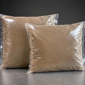 May include: Two square, clear plastic bags filled with a fine, light brown granular substance. The bags are sealed and positioned one slightly behind the other, showcasing the uniform texture and sandy color of the contents inside each package.