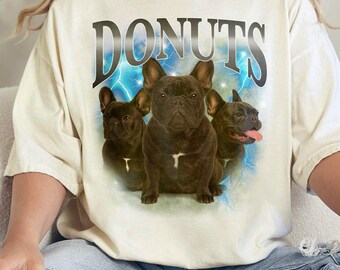 Może przedstawiać: Biały T-shirt z grafiką przedstawiającą trzy buldogi francuskie i napis "DONUTS" w pogrubionym, czarnym foncie. Psy są otoczone niebieskimi błyskawicami i białym tłem chmur.