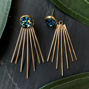 May include: A pair of gold earrings with a blue and green glitter accent. Each earring has a round, glittery charm and seven gold bars extending from the bottom.