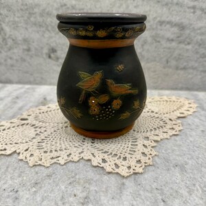 May include: A black ceramic vase with a detailed hand-painted design of two birds and foliage. The vase has a dark brown rim and base, with gold accents. The vase is sitting on a cream-colored lace doily.