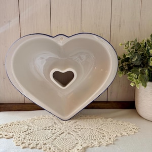 May include: A white heart-shaped ceramic baking dish with a blue rim and a smaller heart-shaped opening in the center. The dish is on a white surface with a cream-colored lace doily. A white pot with greenery is in the background.