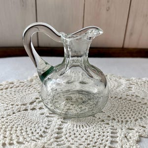 May include: A clear glass pitcher with a curved handle and a small spout. The pitcher has a round body and a green accent at the base of the handle. It sits on a white, crocheted doily against a light wood background.