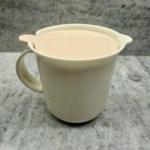 May include: Cream-colored mug with a matching lid. The mug has a handle and a spout. The lid is a light peach color and fits snugly on top. The mug is made of a smooth, solid material.