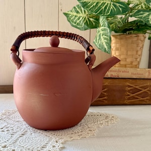 以下が含まれることがあります： テラコッタ色のティーポット。編み込みの取っ手と、お揃いの蓋が付いています。ティーポットは丸みを帯びたボディと注ぎ口があります。取っ手は茶色の編み込み素材です。ティーポットは白いレースのドイリーの上に置かれています。