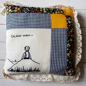 May include: A square patchwork pillow featuring a cream-colored center panel with a black line drawing of a person sitting on a hill and the text "OH, DEAR WORLD-". The pillow includes black and white gingham, yellow, and floral fabric, with lace trim.
