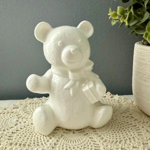 Può includere: Ornamento a forma di orsetto in ceramica bianca con fiocco e piccolo regalo quadrato. L'orsetto è appoggiato su un centrino all'uncinetto color crema. Sullo sfondo c'è una pianta in un vaso bianco. Il centrino misura circa 30 cm di diametro.