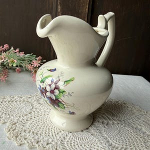 May include: Cream-colored ceramic pitcher with a flared spout and handle. The pitcher features a floral design with purple and white flowers and green leaves. The pitcher sits on a pedestal base.