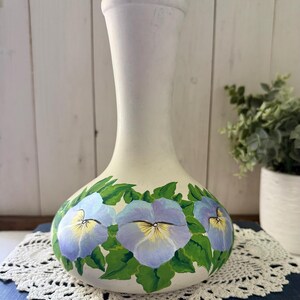 Könnte beinhalten: Eine matte weiße Keramikvase mit einem schmalen Hals und einem abgerundeten Boden. Der untere Teil der Vase ist handbemalt mit einem durchgehenden Band aus hellvioletten und blauen Stiefmütterchen mit gelben Mitten und grünen Blättern.
