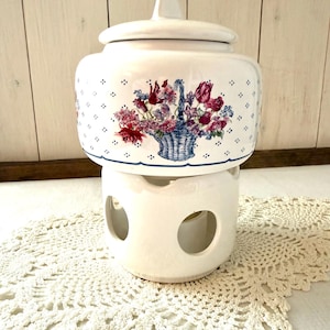 Può includere: Vaso in ceramica bianca con coperchio, con un design a cestino floreale in rosso, viola e blu. Il vaso poggia su uno scaldavivande abbinato con aperture circolari. Il design include piccoli accenti floreali blu.