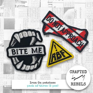 Puede incluir: Conjunto de tres parches termoadhesivos. Incluye un parche de colmillos de vampiro en blanco y negro con el texto "BITE ME", un parche rojo y blanco "DO NOT APPROACH" y un parche triangular amarillo con el texto "RBF".