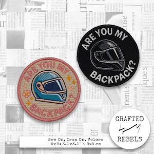 Puede incluir: Dos parches circulares con el texto "ARE YOU MY BACKPACK?" y un gráfico de casco. Un parche es negro con texto blanco y un gráfico de casco, el otro es beige con un gráfico de casco azul. Los parches miden aproximadamente 8 cm de diámetro.