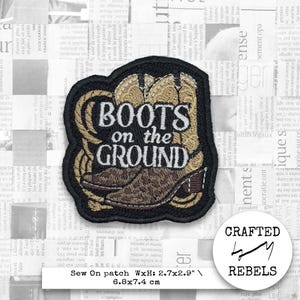 Puede incluir: Un parche bordado marrón y beige con un par de botas de vaquero y el texto "BOOTS on the GROUND". El parche está rodeado por un borde negro y tiene un fondo blanco. El parche mide 6,8 cm por 7,4 cm.