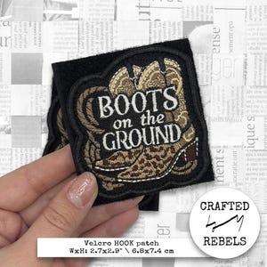 Könnte beinhalten: Ein gestickter Patch in Schwarz und Braun mit einem Paar Cowboystiefeln und dem Text "BOOTS on the GROUND". Der Patch hat einen Klettverschluss zum Anbringen an Kleidung oder Taschen. Der Patch misst 6,8 cm x 7,4 cm.